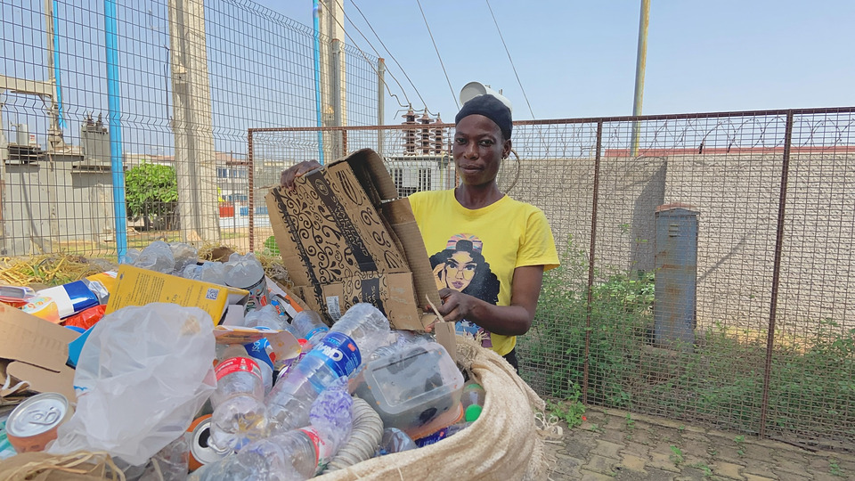 A woman holding flattened cardboard beside a sack of sorted plastic bottles at a collection yard.
